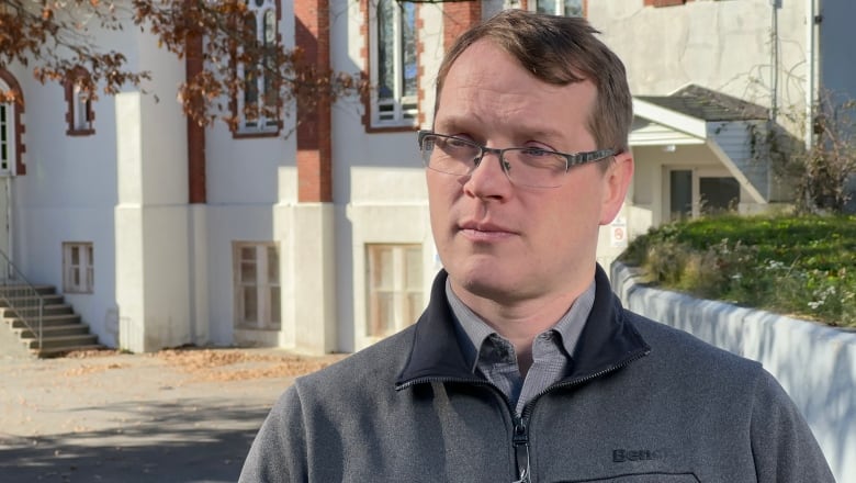 A white man with glasses and brown hair wearing a grey sweater stands outside with a large white building behind him