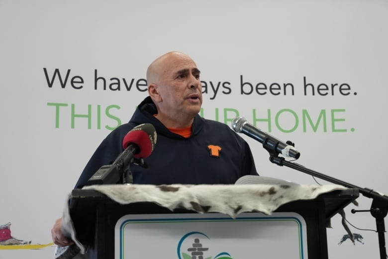 A bald man stands at a podium. He's wearing an orange t-shirt along with a pin of an orange shirt over his heart.